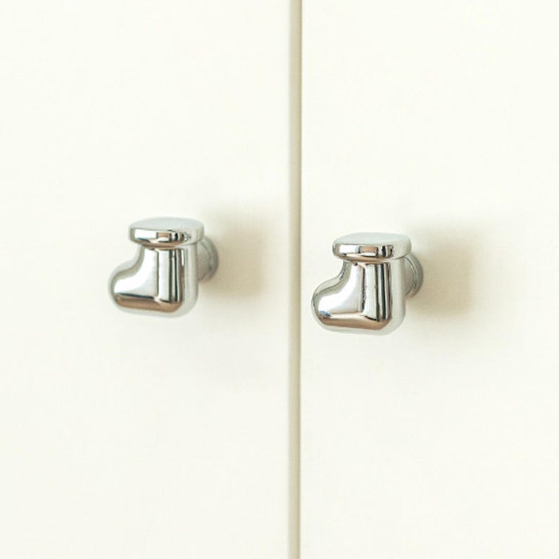 Pu&ograve; includere: Due pomelli cromati lucidi a forma di calzino sono montati su un'anta di un armadio color crema. I pomelli hanno una superficie liscia e riflettente, che aggiunge un tocco moderno al mobile.