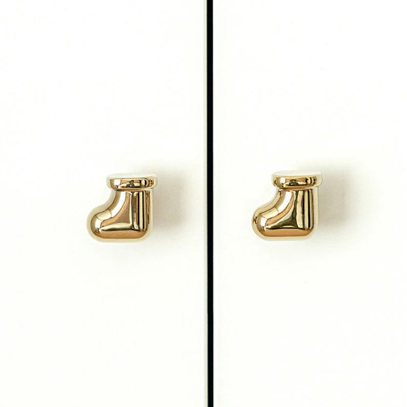 Pu&ograve; includere: Due pomelli dorati per armadi a forma di calza natalizia sono montati su un'anta bianca. I pomelli sono lucidi e riflettono la luce, aggiungendo un tocco di eleganza all'armadio.