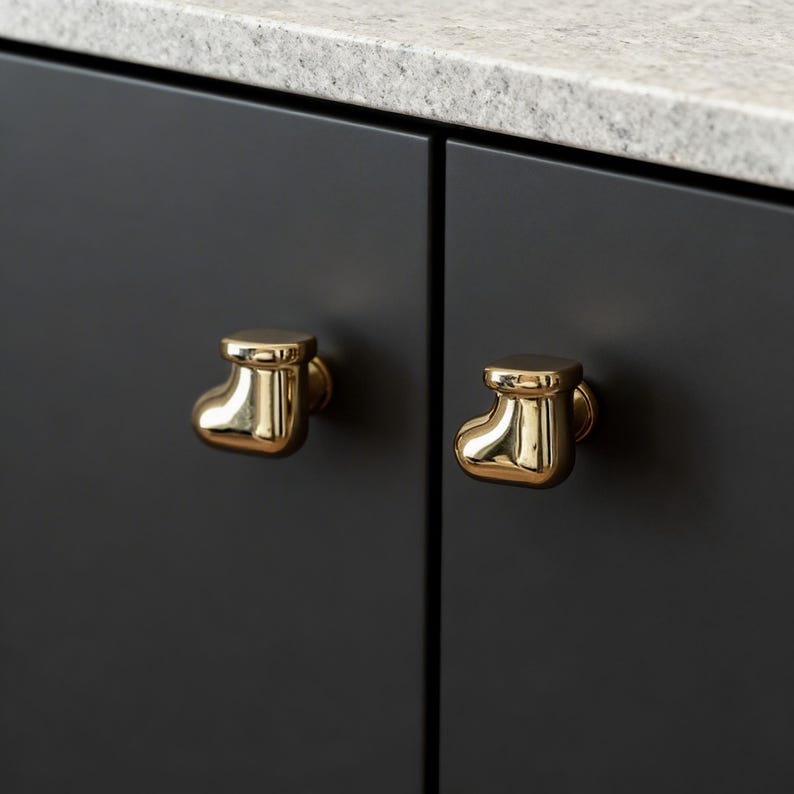 Pu&ograve; includere: Due pomelli dorati per armadi a forma di stivaletto da bambino sono montati su un'anta di un armadio grigio scuro. I pomelli hanno una superficie lucida e riflettente. Sullo sfondo &egrave; visibile un piano di lavoro colorato.