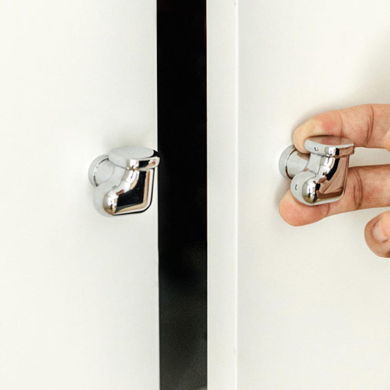 Pu&ograve; includere: Primo piano di due pomelli cromati per armadi. Uno &egrave; attaccato a una porta bianca, l'altro &egrave; tenuto da una mano. I pomelli hanno una parte superiore arrotondata e una base curva e angolare. Sullo sfondo &egrave; visibile una striscia nera.