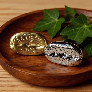 Könnte beinhalten: Zwei dekorative Objekte in Form von Kaffeebohnen, eines goldfarben und eines silberfarben, liegen auf einem Holztablett. Die goldene Bohne hat eine strukturierte Oberfläche, während die silberne das Licht reflektiert. Grüne Blätter sorgen für einen natürlichen Touch.