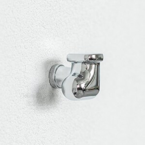 Pu&ograve; includere: Un raccordo idraulico cromato, progettato per il montaggio a parete. Il raccordo presenta un corpo curvo con una parte superiore rettangolare e una base circolare. Probabilmente fa parte di un sistema di approvvigionamento idrico.