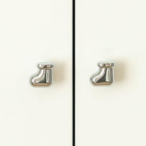 Pu&ograve; includere: Due pomelli per armadietti cromati lucidi a forma di calza natalizia. I pomelli sono montati su un'anta bianca. I pomelli sono riflettenti e hanno una superficie liscia e lucida.