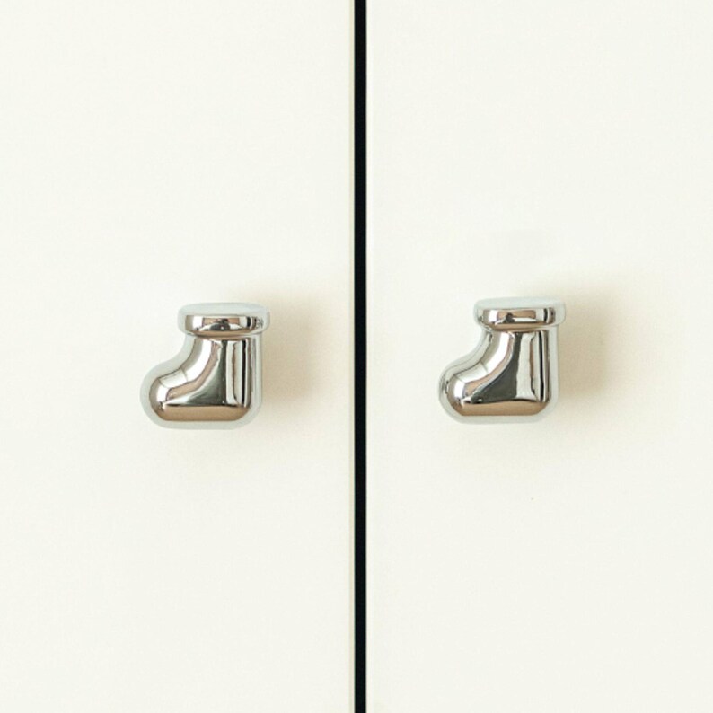 Pu&ograve; includere: Due pomelli per armadietti cromati lucidi a forma di calza natalizia. I pomelli sono montati su un'anta bianca. I pomelli sono riflettenti e hanno una superficie liscia e lucida.