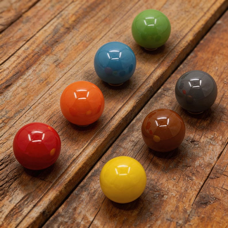 Puede incluir: Un conjunto de siete pomos de cer&aacute;mica brillantes en varios colores: rojo, naranja, amarillo, verde, azul, marr&oacute;n y gris. Cada pomo tiene una hendidura cuadrada. Los pomos est&aacute;n colocados sobre una superficie de madera r&uacute;stica.