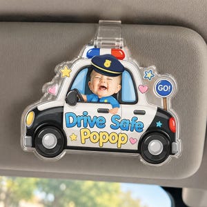 May include: A clear plastic car visor clip featuring a cartoon police car with a baby dressed as a police officer. The car is white and black with blue text that reads "Drive Safe Popop". The baby is smiling and wearing a police hat.