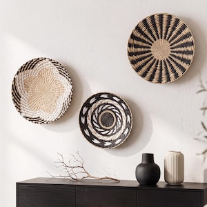 May include: Three circular woven wall baskets featuring intricate patterns in black, white, and natural tan fibers. The baskets are displayed in a cluster on a wall above a dark wooden cabinet, which holds a black vase and a ribbed cream-colored vase.