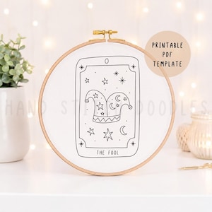 May include: A circular wooden embroidery hoop containing white fabric with a black line art design of a tarot card titled The Fool. The design features a jester hat surrounded by stars and crescent moons, with the text PRINTABLE PDF TEMPLATE.
