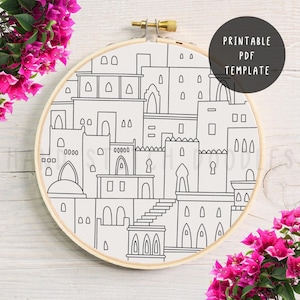 May include: A black and white embroidery hoop featuring a line drawing of a cityscape with arches, windows, and rooftops. The text "PRINTABLE PDF TEMPLATE" is in a black circle. The hoop is set against a white background with pink flowers.