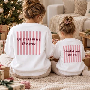 May include: Two white crewneck sweatshirts featuring a pink vertical striped square on the back. The text "Christmas Crew" is printed in dark red lettering across the center of the stripes on both garments, which are worn by two individuals.