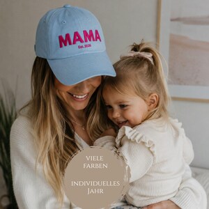 May include: A light blue baseball cap featuring the word MAMA embroidered in bold pink letters, with the text Est. 2026 embroidered in smaller pink letters underneath. The cap has a structured crown, a curved brim, and a button on the top.