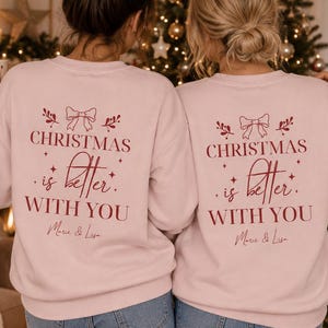 May include: Two light pink crewneck sweatshirts featuring a red graphic on the back. The design includes a bow, sprigs, and the text "CHRISTMAS is better WITH YOU" followed by the names "Marie & Lisa" in a cursive script font.