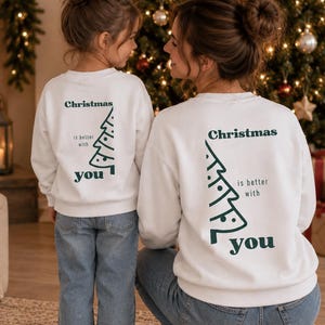 May include: Two white crewneck sweatshirts featuring a dark green graphic on the back. The design includes a stylized Christmas tree outline and the text "Christmas is better with you" printed in a dark green serif font on both garments.