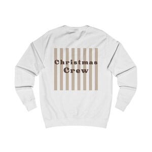 Puede incluir: Una sudadera blanca de manga larga con un gr&aacute;fico central de rayas verticales de color beige. Sobre las rayas aparece el texto "Christmas Crew" en una fuente serif redondeada, negrita y de color marr&oacute;n oscuro, en la espalda.