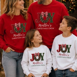 May include: A collection of matching long-sleeve sweatshirts in red and white. Each features the text JOY with a wreath replacing the letter O, topped with a red and white checkered bow. Below, the text reads M&auml;ller's CHRISTMAS CREW.