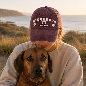 May include: A maroon baseball cap with the text "RIDGEBACK DOG MOM" in white, along with paw prints and a bow. The cap is worn by a person holding a brown dog with a leather collar. The background shows a beach and ocean.