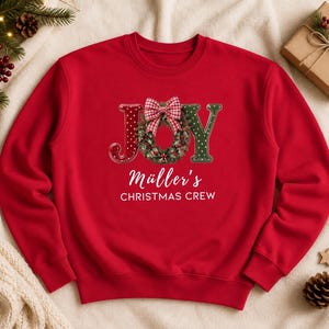 May include: A red crewneck sweatshirt featuring the word JOY in large letters, with the letter O replaced by a green Christmas wreath adorned with a red and white checkered bow. Below the graphic, the text reads M&uuml;ller's CHRISTMAS CREW.