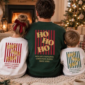 May include: A dark green sweatshirt and two white sweatshirts featuring a red or green vertical striped graphic with gold HO HO HO text. The dark green shirt reads MÜLLER'S FAVOURITE CHRISTMAS BUDDY SINCE 2020, while the white shirts read DADDY'S FAVOURITE CHRISTMAS BUDDY SINCE 2020 or 2024.