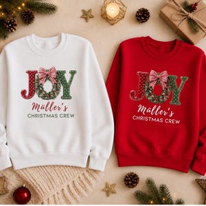 May include: A white sweatshirt and a red sweatshirt, each featuring the text JOY with a wreath replacing the O, a red and white checkered bow, and the printed text M&uuml;ller's CHRISTMAS CREW in a script and sans-serif font combination.