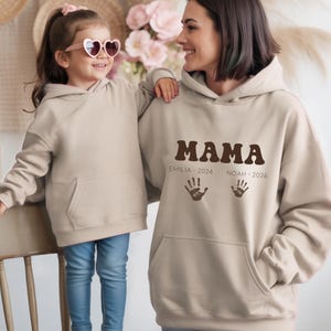 Puede incluir: Dos sudaderas con capucha beige, una usada por una mujer y la otra por una niña. La sudadera de la mujer dice "MAMA" en letras marrones, con "EMILIA 2024" y "NOAH 2026" debajo. La niña lleva gafas de sol en forma de corazón y vaqueros azules.