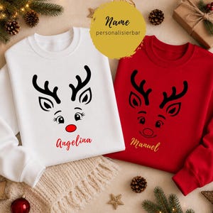 Puede incluir: Una sudadera blanca y una sudadera roja, cada una con un gráfico negro de una cara de reno. La sudadera blanca tiene una nariz roja y el nombre Angelina en rojo. La sudadera roja lleva el nombre Manuel escrito en color dorado.