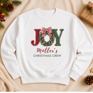 May include: A white long-sleeve sweatshirt featuring the word JOY in large letters, where the O is replaced by a green Christmas wreath with a red and white gingham bow. Below, the text reads M&uuml;ller's CHRISTMAS CREW in red and dark green.