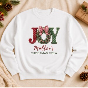 May include: A white crewneck sweatshirt featuring a graphic with the word JOY, where the O is replaced by a green wreath with a red and white gingham bow. Below the graphic, the text reads M&uuml;ller's CHRISTMAS CREW in red and dark green.