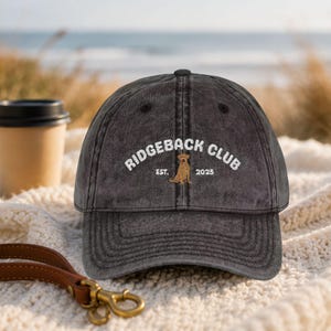 May include: A charcoal gray baseball cap with the text "RIDGEBACK CLUB" embroidered in white, along with a dog graphic and the year "2025". The cap has a curved brim and a distressed look, suggesting a vintage style.