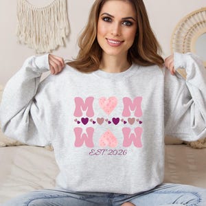 Puede incluir: Sudadera gris claro con la palabra "MOM" en rosa, con detalles de corazones y el texto "EST. 2026". La sudadera es de cuello redondo.