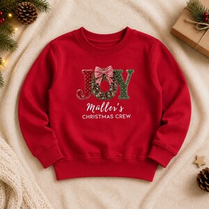 May include: A red long-sleeve sweatshirt featuring the word JOY with a green wreath replacing the letter O, topped with a red and white checkered bow. Below the graphic, the text reads M&uuml;ller's CHRISTMAS CREW in white lettering.