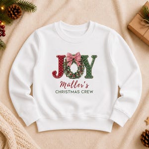 May include: A white long-sleeve sweatshirt featuring the word JOY with a Christmas wreath replacing the letter O. The wreath has a red and white checkered bow. Below the graphic, the text reads M&uuml;ller's CHRISTMAS CREW in red and dark green.