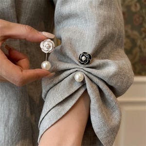 May include: A grey linen-textured sleeve featuring a gathered cuff detail. The sleeve is adorned with two decorative camellia-shaped buttons, one white and one black, each with a central pearl accent, plus a single white pearl button below.