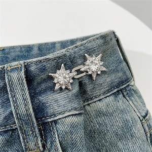 Pode incluir: Um ajustador de cintura de metal prateado com dois ornamentos em forma de estrela decorados com cristais transparentes facetados. As duas estrelas estão ligadas por um pequeno gancho de metal, concebido para ajustar o tecido de ganga azul.