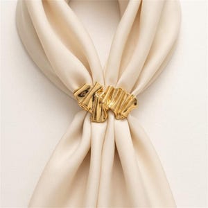 Könnte beinhalten: Ein glänzender goldfarbener Schalring mit einer welligen, organischen und gerippten Textur. Das metallische Accessoire ist mit einem glatten, cremefarbenen Stoffschal durchzogen, der das Material in einer gerafften Anordnung in der Mitte hält.