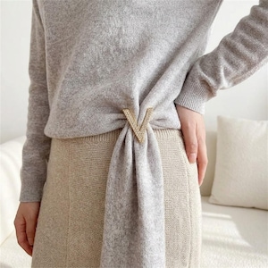 Könnte beinhalten: Ein hellgrauer Langarmpullover mit einem V-förmigen, goldfarbenen Metallclip, der mit kleinen klaren Kristallen besetzt ist. Der Clip rafft den Stoff des Pullovers an der Taille und erzeugt einen drapierten Effekt über einem beigen Rock.