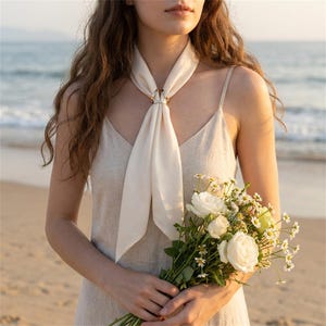May include: A cream-colored scarf secured with a metallic gold-toned ring, worn with a sleeveless cream-colored dress. The scarf is tied in a simple knot at the neck, and the outfit is paired with a bouquet of white roses and small daisies.