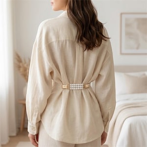 Puede incluir: Una camisa beige de manga larga con botones, ajustada en la espalda con un clip de metal dorado que presenta tres filas horizontales de perlas blancas. La camisa tiene un corte holgado, cuello y puños abotonados, creando un efecto fruncido.