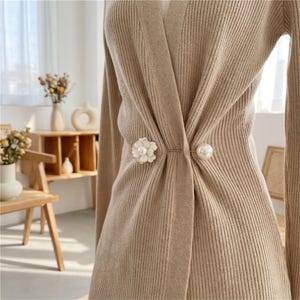 Op de afbeelding: Een beige vest van geribd breiwerk met lange mouwen en een sluiting die wordt vastgezet met een decoratieve metalen speld. De speld bestaat uit een wit bloemvormig accent aan de ene kant en een gladde, ronde witte parel aan de andere kant.