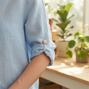 May include: A light blue linen-textured shirt with a rolled-up sleeve secured by a smooth, beige, circular-topped cuff accessory. The fabric has a visible weave, and the sleeve is folded neatly above the elbow, held in place by the fastener.