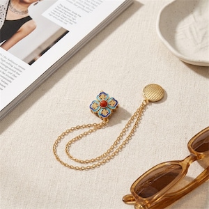 Puede incluir: Un broche dorado con forma de trébol de cuatro hojas, detalles en esmalte azul y una cuenta roja central. El broche incluye dos delicadas cadenas doradas conectadas a un disco circular dorado con textura en el extremo opuesto de las cadenas.