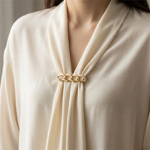 Puede incluir: Una blusa de color crema con escote en V fruncido en el centro por un accesorio de metal dorado con forma de eslabones de cadena. La tela tiene una textura suave y lisa con pliegues verticales que caen desde el escote, creando un efecto drapeado.