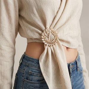Può includere: Una spilla o clip circolare dorata per foulard con una corona di cristalli trasparenti a taglio marquise. L'accessorio è fissato sulla parte anteriore di un top a portafoglio color beige con texture in lino, raccogliendo il tessuto.