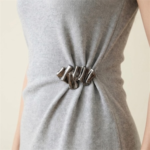 Könnte beinhalten: Ein ärmelloses Strickkleid in Hellgrau mit einer metallischen, dunkel silberfarbenen Brosche an der Taille. Die Brosche hat eine abstrakte, gewellte und organische Form, die den Stoff des Kleides in weichen, vertikalen Falten an der Seite rafft.