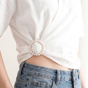 Puede incluir: Una camisa blanca de manga corta con una hebilla circular de perlas de imitación en la cintura que frunce la tela. La camisa se combina con unos vaqueros azules. El accesorio de perlas es redondo y consta de pequeñas cuentas blancas.