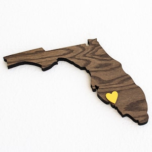 May include: A wooden cutout of the state of Florida with a yellow heart marking a location. The wood has a dark brown grain.