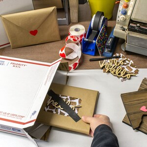 May include: A white Priority Mail box with a brown package inside. The package is wrapped in brown paper and has a black tape seal. The package has a wooden cutout of the word "hello" on it. There are two wooden state shapes with pink hearts on them.