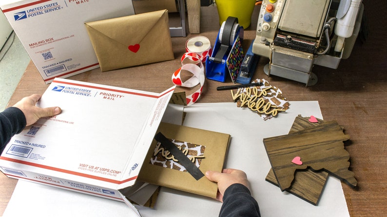 May include: A white Priority Mail box with a brown package inside. The package is wrapped in brown paper and has a black tape seal. The package has a wooden cutout of the word "hello" on it. There are two wooden state shapes with pink hearts on them.