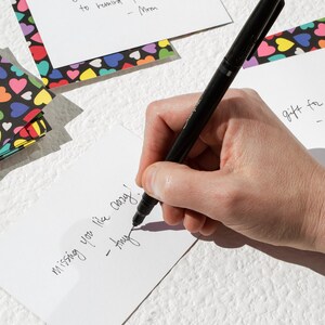 May include: A hand writing a note on a white card with black ink. The note says "missing you like crazy! - hug". The card is surrounded by colorful heart-patterned paper.