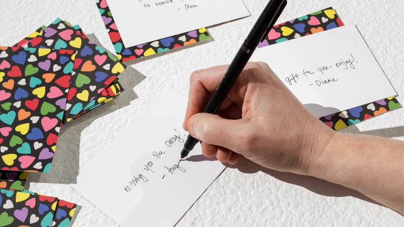 May include: A hand writing a note on a white card with black ink. The note says "missing you like crazy! - hug". The card is surrounded by colorful heart-patterned paper.