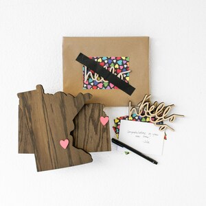 May include: A brown wooden cutout of the state of Minnesota with two pink hearts attached. A brown paper envelope with a black tape seal and a colorful heart pattern inside. A white card with the word "hello" written on it. A black pen and a white card with a handwritten message.